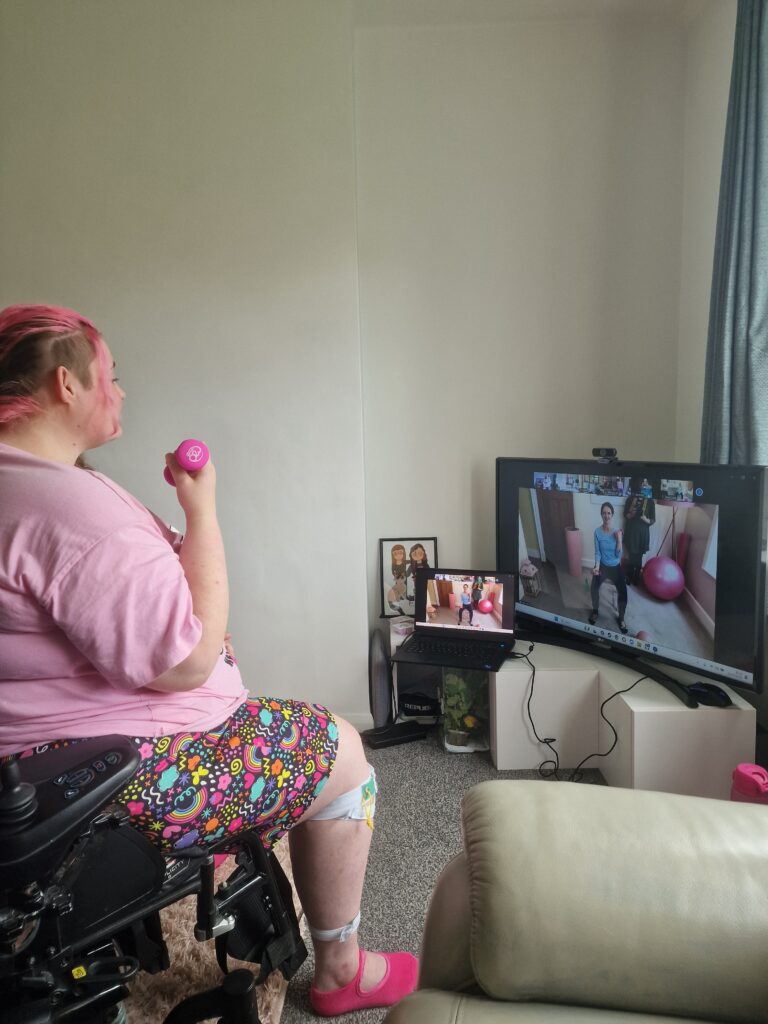 One of our members Sam sitting in her wheelchair in a strong position holding pink weights. On the screen in front of her is our teacher Kerry demonstrating a move.