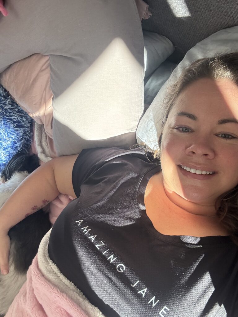 Leanne lying in bed smiling with her hand resting on Stanley, her white and grey Shih Tzu. She is under a pink blanket and lying on grey pillows. her tshirt is black with the words Amazing Jane on the front.
