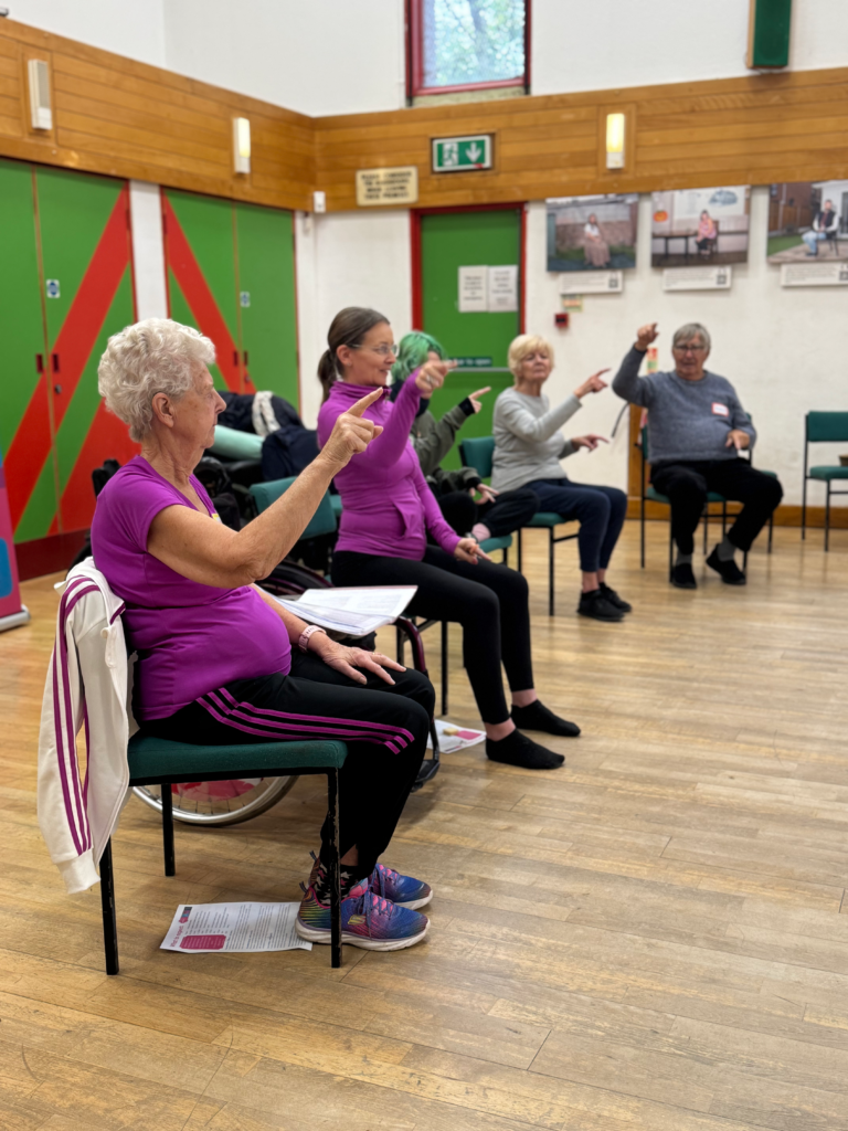 Suzanne wearing a deep pink top and black leggings, sitting on a chair, teaching love to move at the Devon Accessible Exercise Showcase